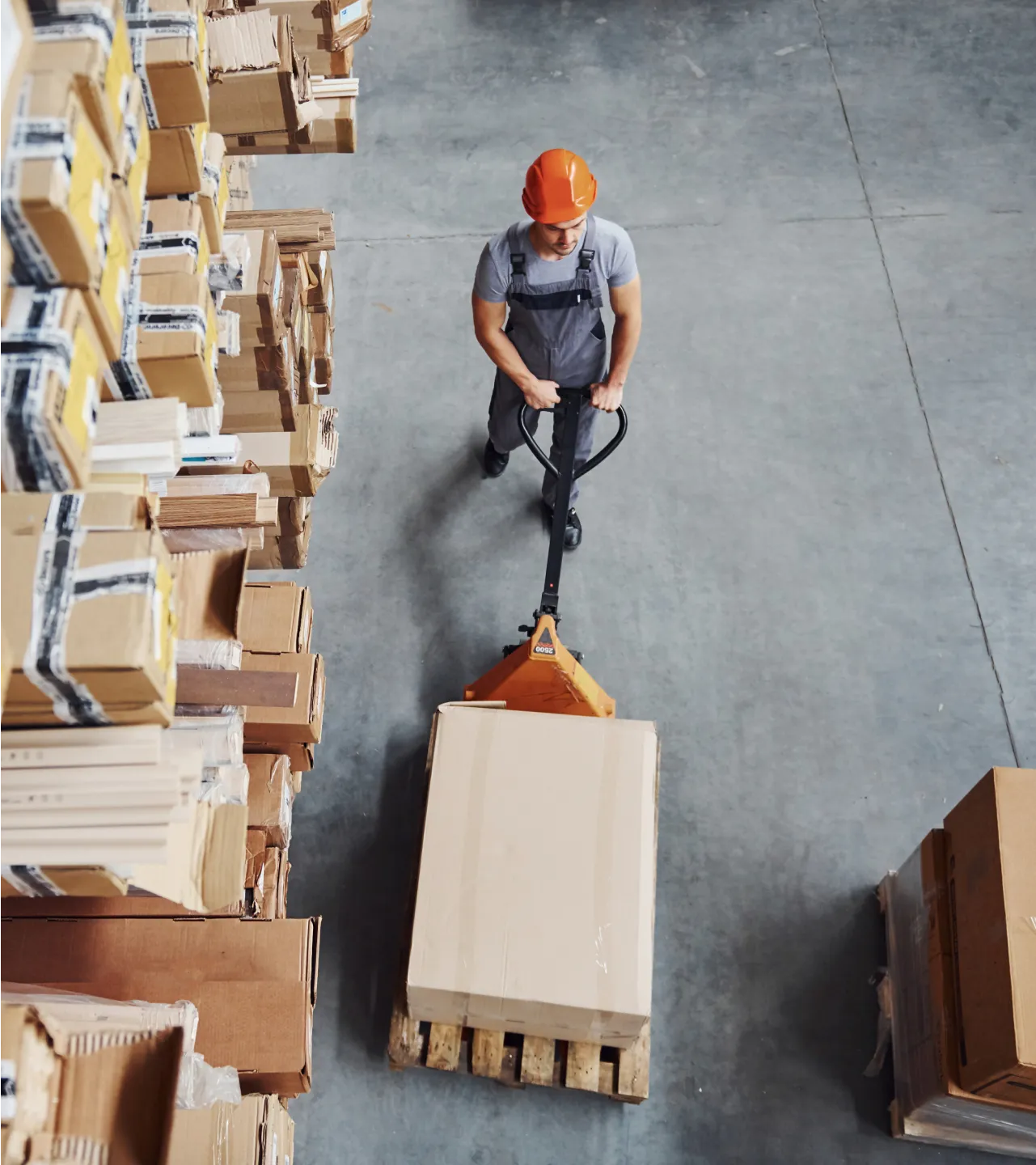 Worker in a warehouse pushing a pallet jack loaded with a large cardboard box between tall shelves of inventory.