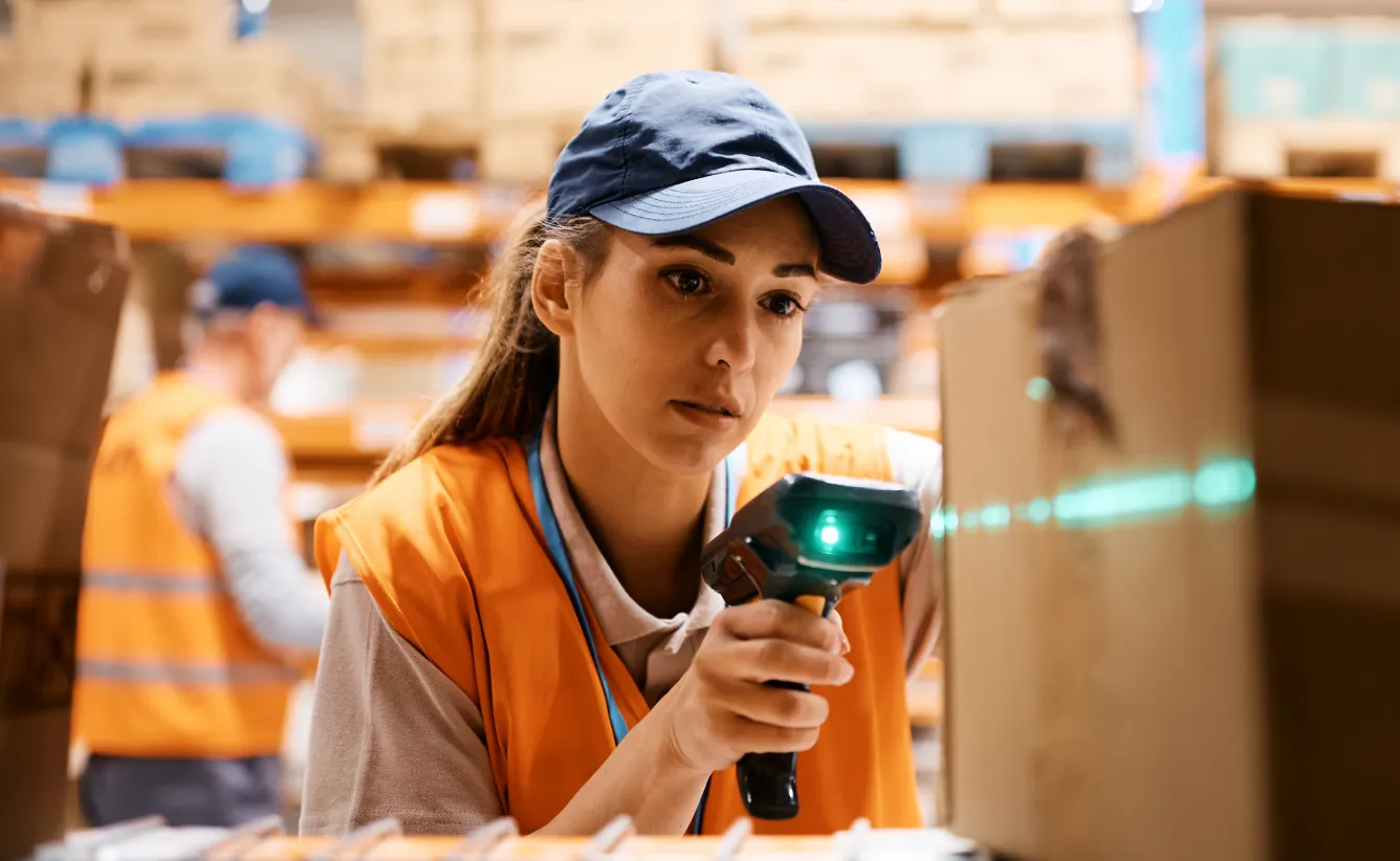 A female warehouse worker scans a barcode on a box using a handheld scanner.