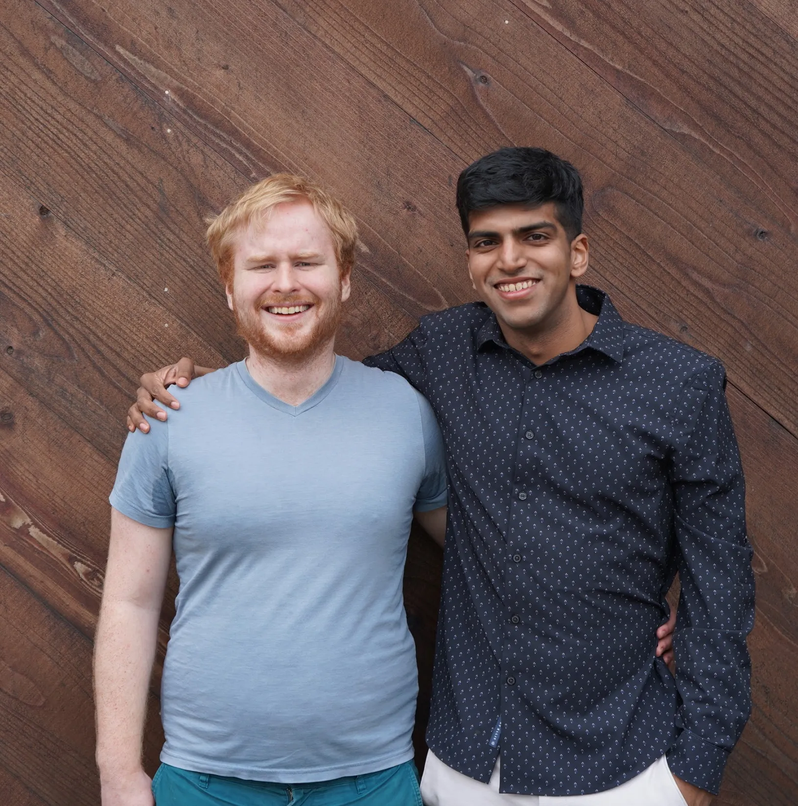 Two men, one with red hair and a beard in a light blue t-shirt, and the other with short dark hair in a dark blue shirt, are smiling with their arms around each other in front of a wooden wall.