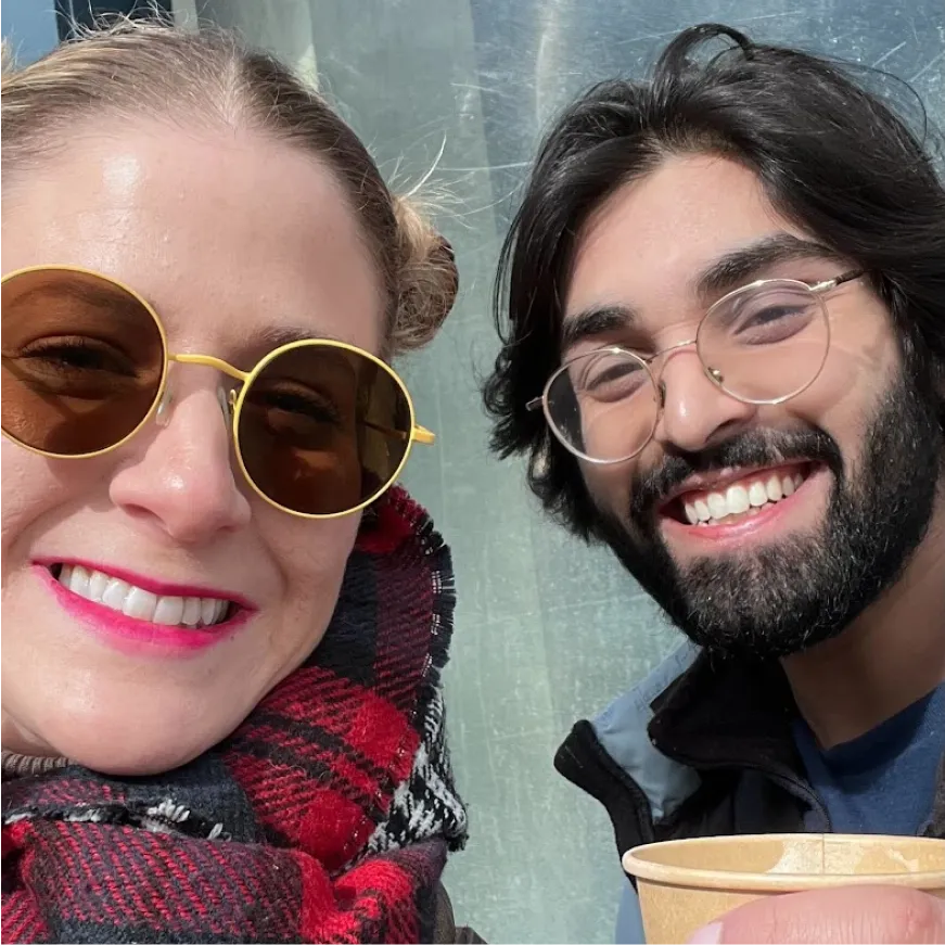 Two team members smiling at the camera, one wearing round sunglasses and a red checkered scarf, the other with glasses and a beard, holding a cup.