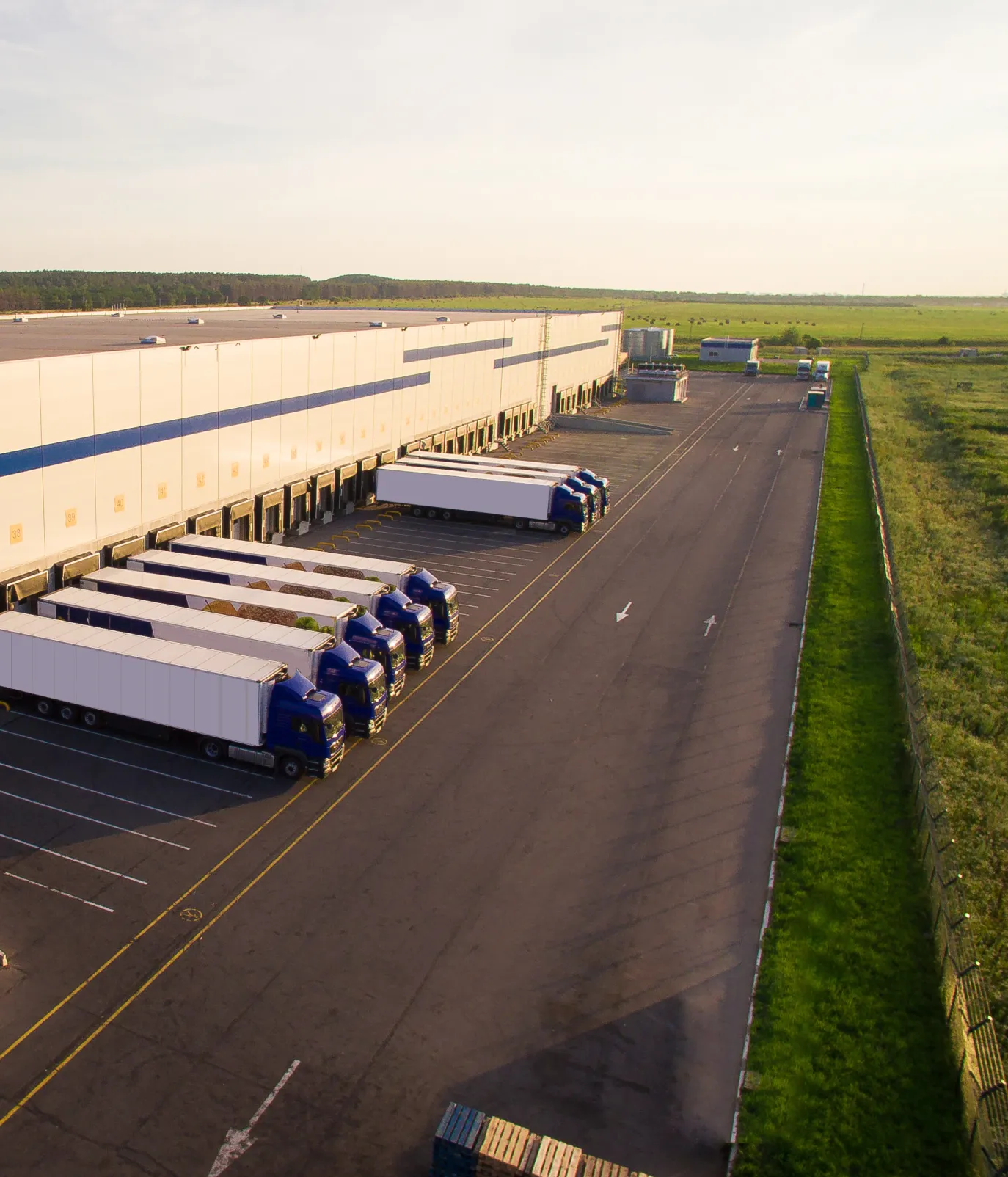 Aerial view of a distribution warehouse with rows of trucks parked at loading docks, surrounded by green fields under a clear sky.