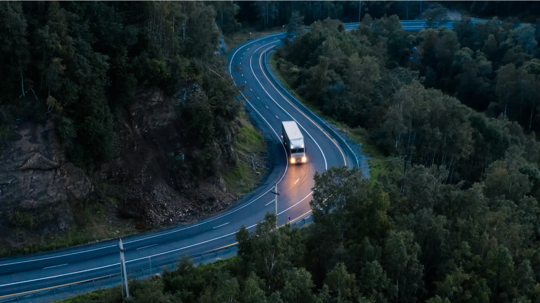 An aerial view shows a truck driving on a winding road through a forested area at dusk. The road curves sharply, surrounded by dense trees.