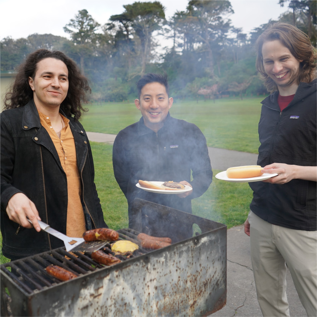 Three men, one with long hair in a black jacket, one with short dark hair in a black jacket, and one with long blonde hair in a black jacket, are smiling while grilling food at a park.