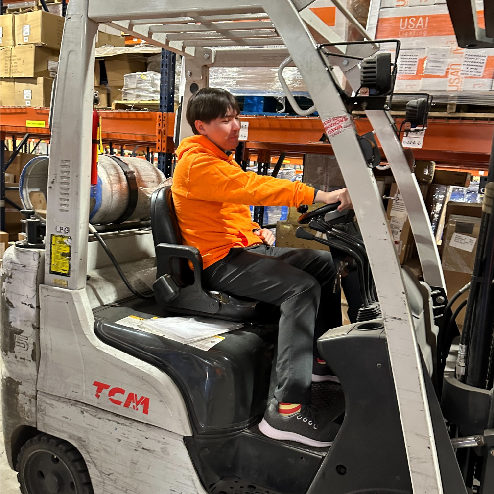 A man with short brown hair wearing an orange sweater is sitting in a white forklift inside a warehouse.