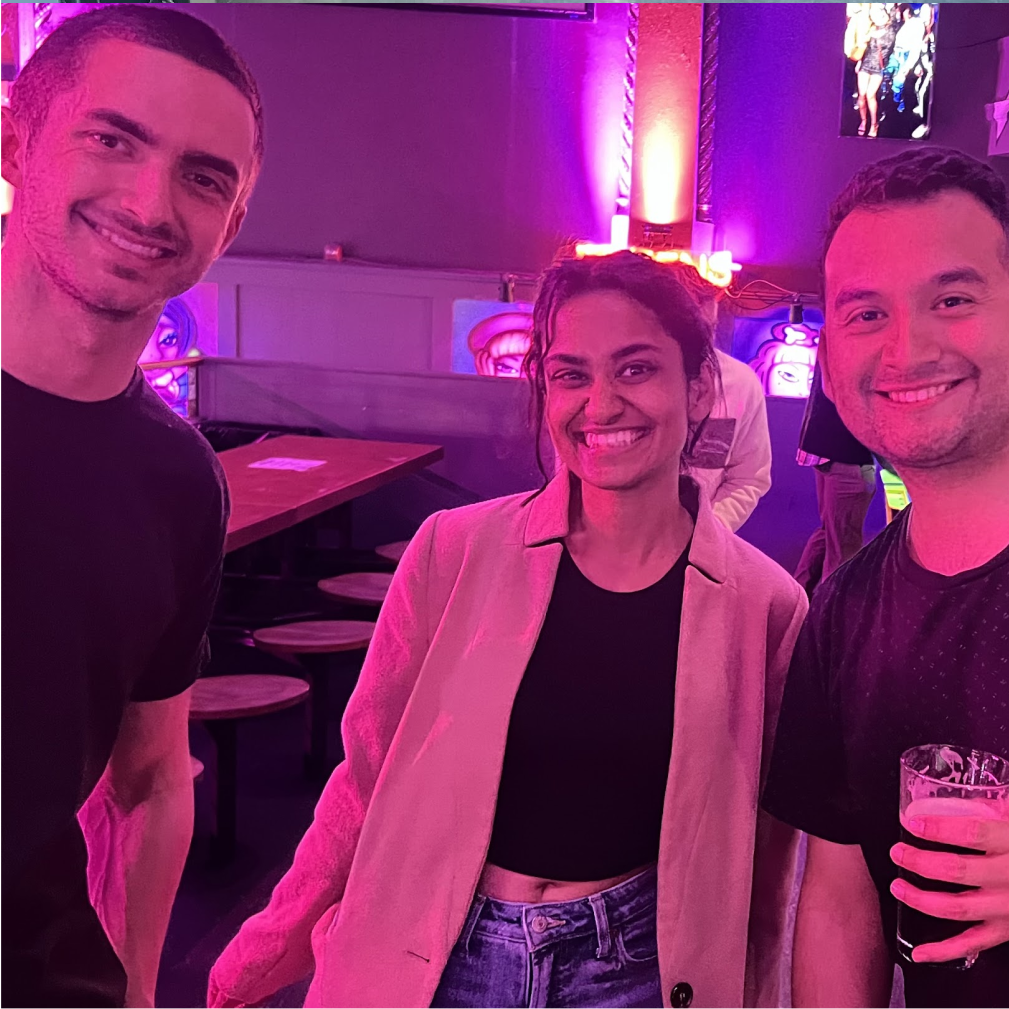 Three people, two men in black t-shirts and a woman in a light blazer, are smiling together in a dimly lit room with purple lighting.