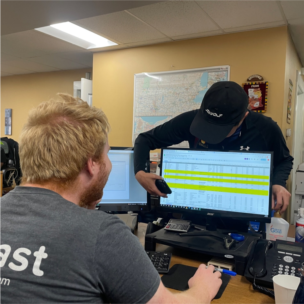 Two men are working at a desk with computer screens; one is seated and looking at the screens, while the other is standing and pointing at the screen.