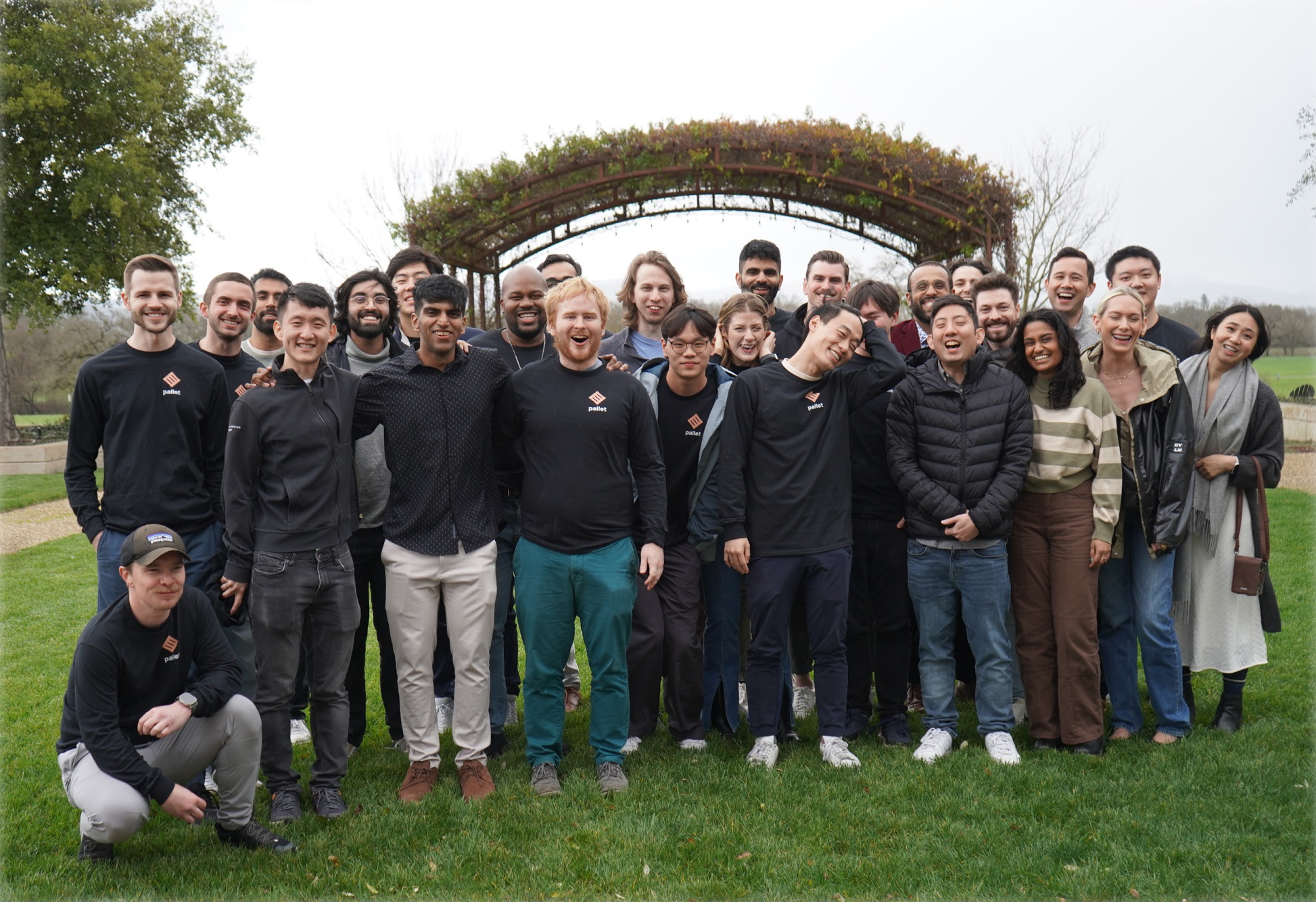 A large group of people are smiling together in an outdoor setting with greenery and a vine-covered archway.